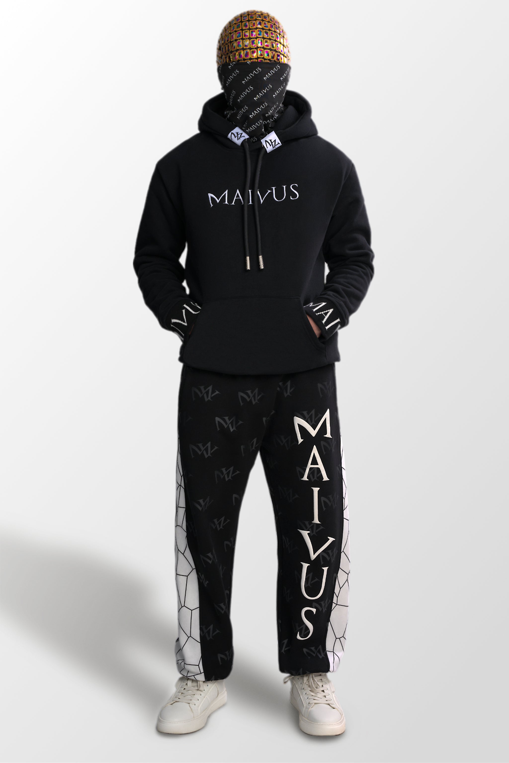 Model wearing a luxury black and white hoodie and black and white joggers. 'MV' branding on joggers with geometric shapes and white embroidery with branded snood on hoodie from 'MAIVUS' against a white studio backdrop