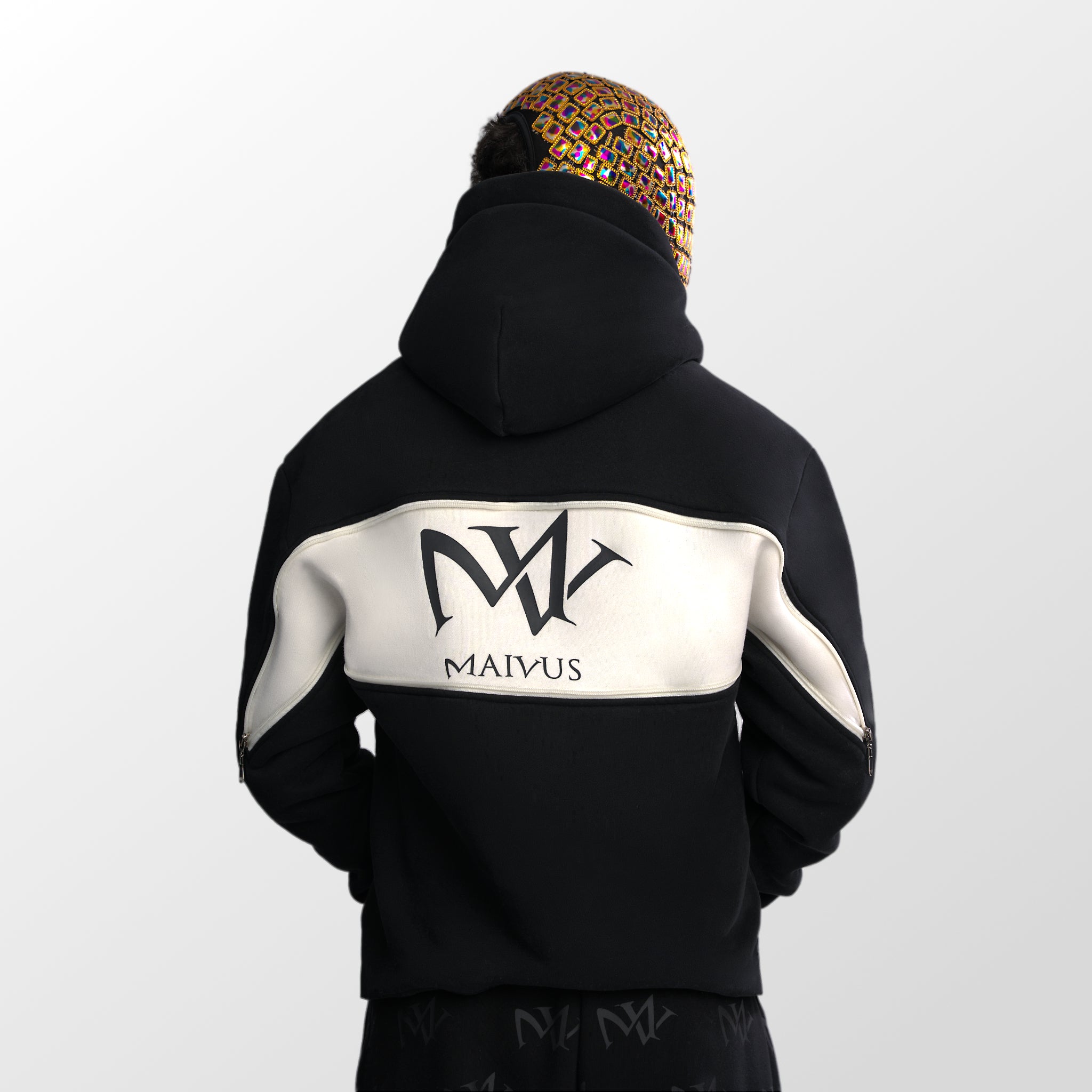 Model wearing a black and white luxury hoodie. Large 'MV' logo on back of hoodie from 'MAIVUS' against a white studio backdrop