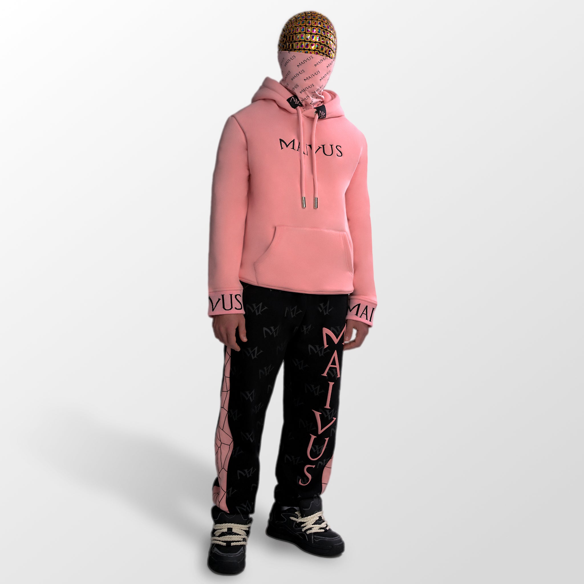Model wearing a luxury pink and black hoodie and black and pink joggers. 'MV' branding on joggers with geometric shapes and black embroidery with branded snood on hoodie from 'MAIVUS' against a white studio backdrop
