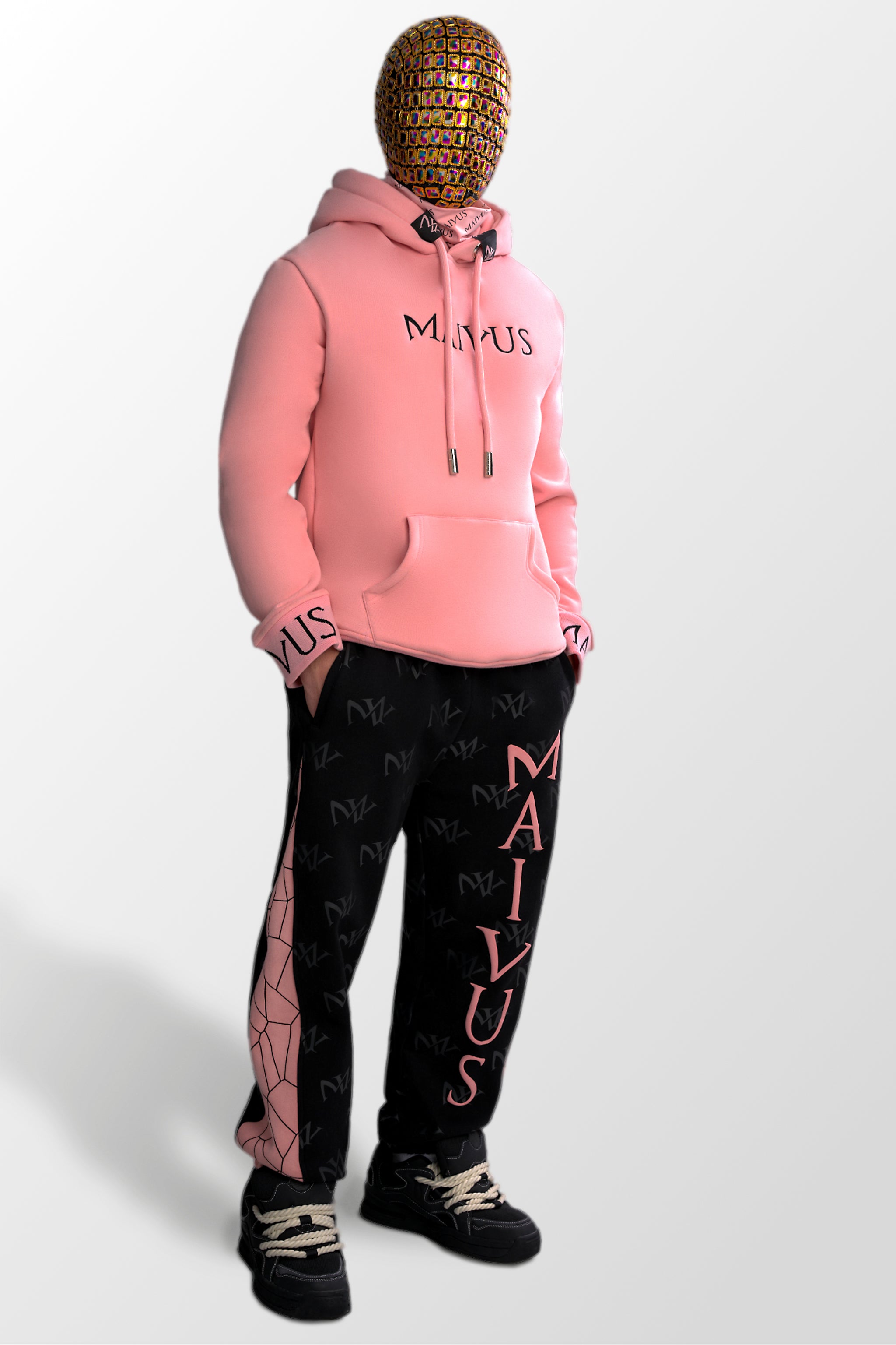 Model wearing a luxury pink and black hoodie and pink and black joggers. 'MV' branding on joggers with geometric shapes and black embroidery with branded snood on hoodie from 'MAIVUS' against a white studio backdrop