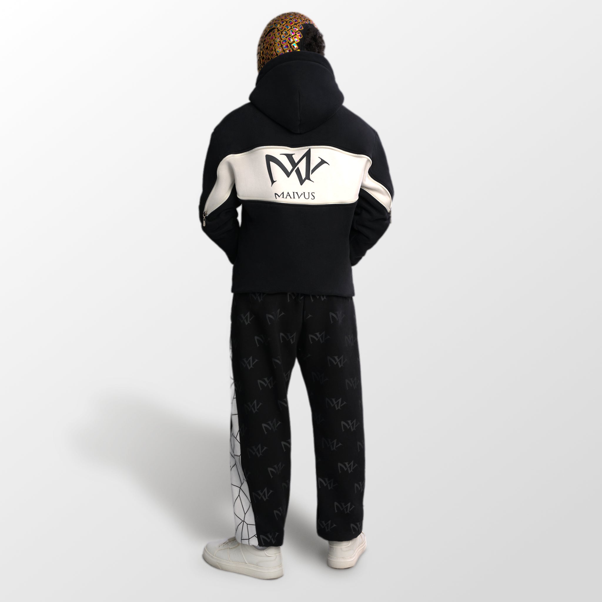 Model wearing a black and white luxury hoodie and joggers. 'MV' branding all over the joggers and large 'MV' logo on back of hoodie from 'MAIVUS' against a white studio backdrop