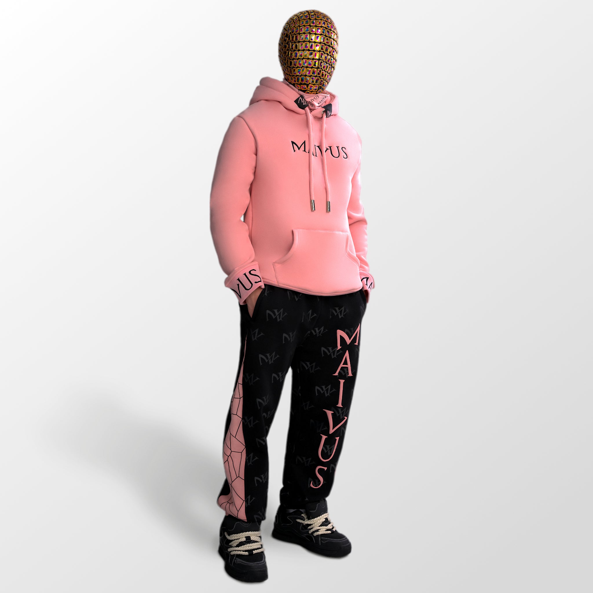 Model wearing a luxury pink and black hoodie and black and pink joggers. 'MV' branding on joggers with geometric shapes and black embroidery on hoodie from 'MAIVUS' against a white studio backdrop