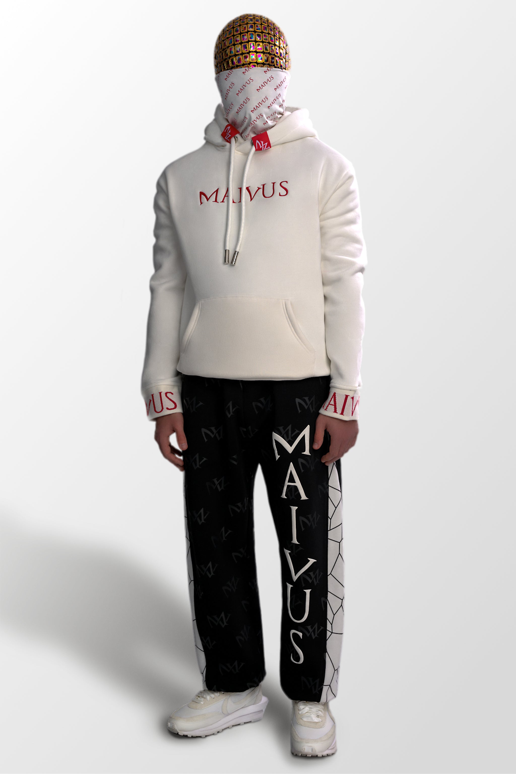 Model wearing a luxury white and red hoodie and black and white joggers. 'MV' branding on joggers with geometric shapes and red embroidery with branded snood on hoodie from 'MAIVUS' against a white studio backdrop